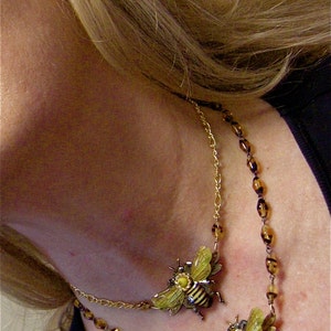 May include: A gold chain necklace with brown beads and two bee charms. The bee charms are made of metal and have yellow and black wings.