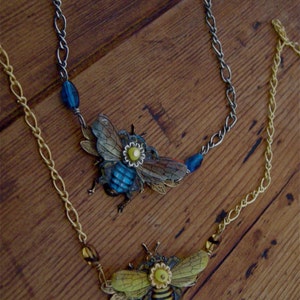 May include: Two necklaces with bee charms. One necklace has a blue bee charm with yellow accents and the other has a yellow and black bee charm with yellow accents. Both necklaces have silver chains and blue and yellow beads.