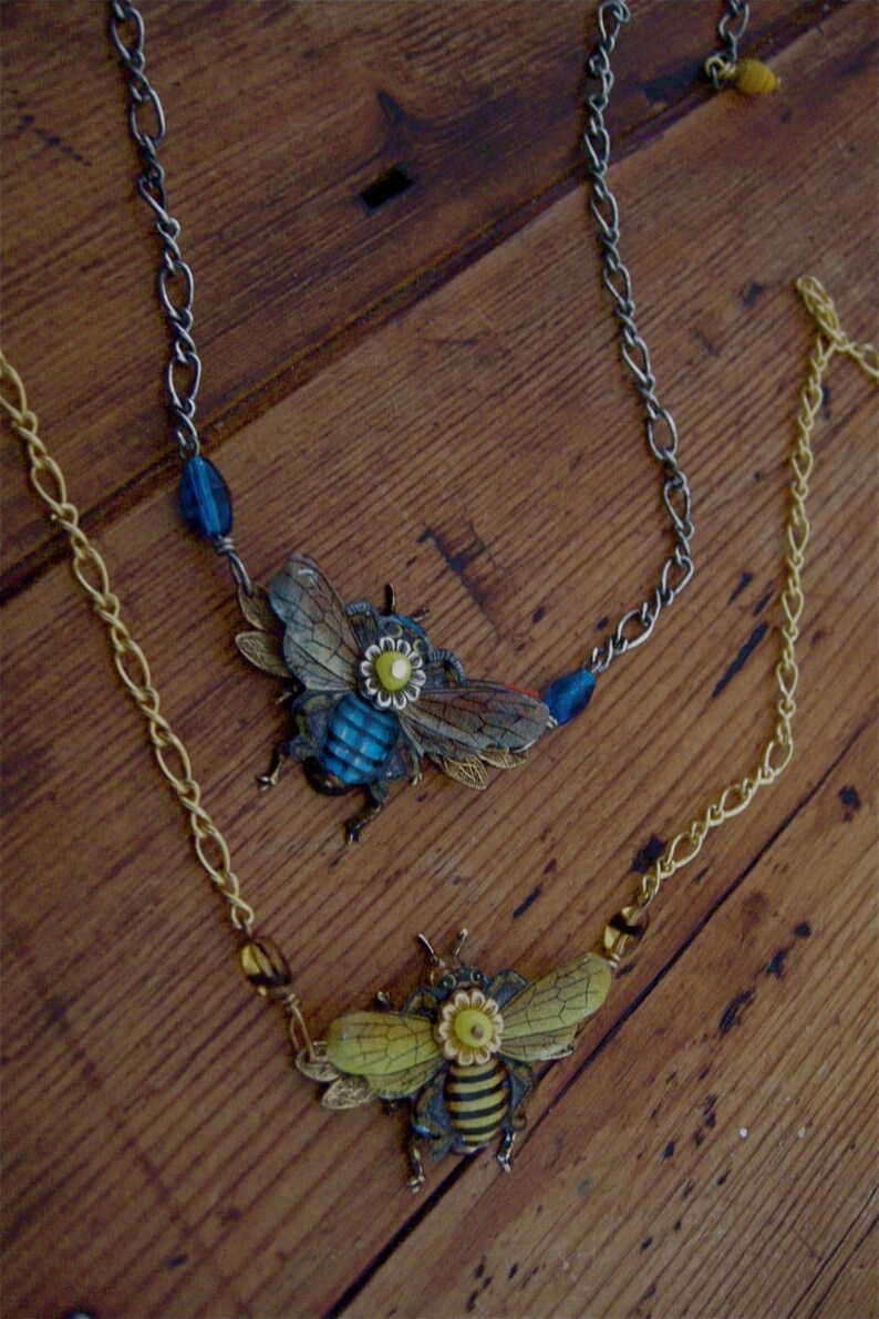 May include: Two necklaces with bee charms. One necklace has a blue bee charm with yellow accents and the other has a yellow and black bee charm with yellow accents. Both necklaces have silver chains and blue and yellow beads.