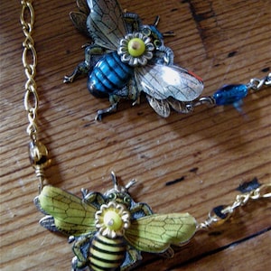 May include: A gold chain necklace with two bee charms. The bees are made of metal and have blue, yellow, and green accents. The bees have wings and are detailed with intricate patterns.