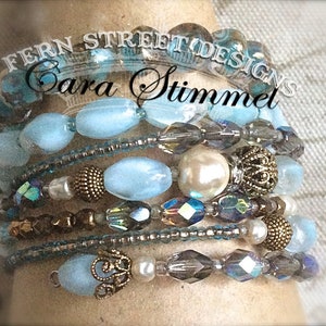 May include: A stack of blue, gray, and white beaded bracelets with gold accents. The bracelets feature a variety of beads, including round, oval, and faceted beads. The text "Fern Street Designs Cara Stimmel" is visible on the bracelets.