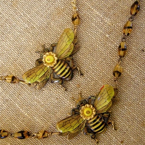 May include: A gold chain necklace with two bee charms. The bees are made of metal and have black and yellow stripes. The bees have yellow flowers on their backs. The chain is made of gold-colored metal and has brown beads.