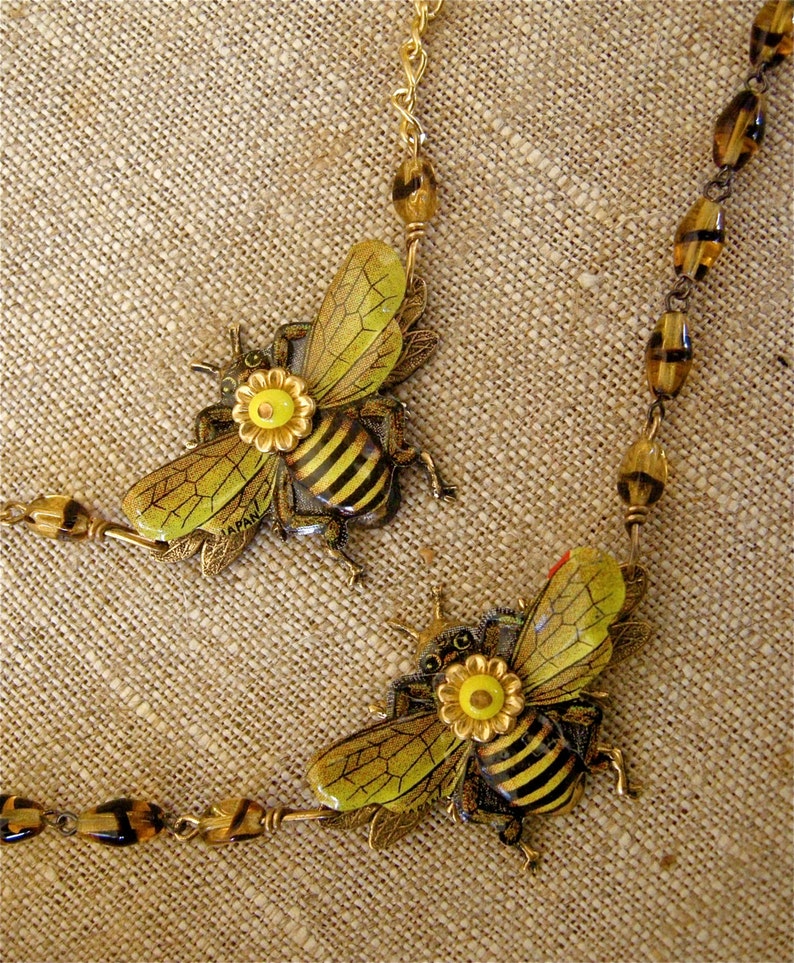 May include: A gold chain necklace with two bee charms. The bees are made of metal and have black and yellow stripes. The bees have yellow flowers on their backs. The chain is made of gold-colored metal and has brown beads.