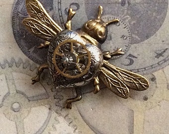 May include: A steampunk-style bee brooch with a heart-shaped body. The bee features brass wings and legs, a silver body with a clockwork design, and a gold-colored gear in the center. The background includes clock faces and the text "Main Street Design Cara Stimmel."
