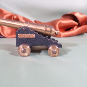 Puede incluir: Un cañón en miniatura con un cañón de latón y ruedas. El carro es azul oscuro con detalles de latón. El fondo presenta tela drapeada en tonos naranja, morado y verde.