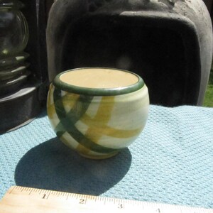 Può includere: Un piccolo vaso in ceramica bianca con strisce verdi e gialle. Il vaso ha un diametro di circa 7,5 cm ed è appoggiato su una tovaglia blu.