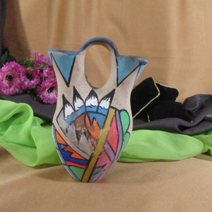 May include: A hand-painted ceramic vase with a geometric design in shades of blue, green, orange, and red. The vase has a unique shape with a wide opening at the top and a narrow base.