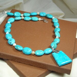 May include: A turquoise and white beaded necklace with a turquoise pendant. The necklace is on a brown surface.