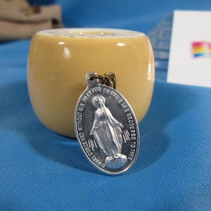 May include: Silver oval-shaped religious medal with a depiction of the Virgin Mary. The medal has an inscription that reads "O Mary conceived without sin pray for us who have recourse to thee".