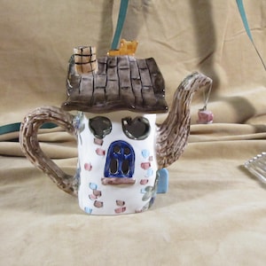 Può includere: Una teiera in ceramica bianca a forma di casa con un tetto marrone, un camino e una finestra blu. La teiera ha un manico marrone e un design floreale rosa e blu. C'è una piccola campana rosa appesa al beccuccio.