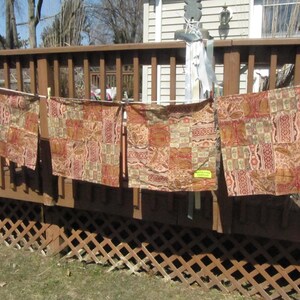 Könnte beinhalten: Vier gemusterte Stoffquadrate hängen an einer Wäscheleine auf einem Holzdeck. Der Stoff ist eine Mischung aus Braun, Rot und Beige mit einem Paisley-Muster.