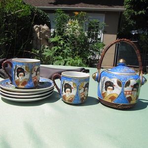 May include: A blue and gold tea set with a teapot and two cups. The teapot has a brown wicker handle and the cups and teapot are decorated with images of women in traditional Japanese clothing.