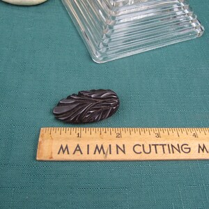 Könnte beinhalten: Eine dunkelbraune, geschnitzte Brosche aus Kunststoff in Form eines Blattes. Die Brosche ist etwa 3,8 cm lang und liegt auf einer grünen Oberfläche neben einem Holzlineal.