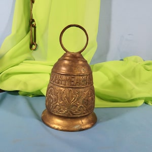 Könnte beinhalten: Eine bronzefarbene Glocke mit Bügelgriff. Die Glocke hat verzierte Blumen- und Rankenmuster und das Wort "REMEMBER" ist oben eingraviert. Die Glocke steht auf einer blauen Oberfläche mit einem grünen Stoffhintergrund.