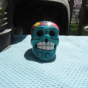 May include: A turquoise ceramic skull with a painted floral design on the top of the head. The skull has white teeth and a black swirl design on the chin.