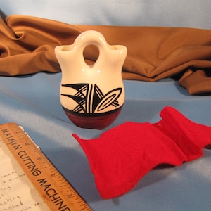 Può includere: Un piccolo vaso in ceramica bianca in stile nativo americano con disegni geometrici neri e rossi. Il vaso ha due aperture nella parte superiore e un manico.