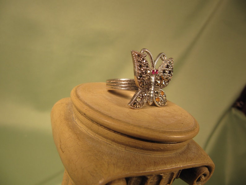 May include: A silver butterfly ring with black and white marquetry and a red gemstone in the center.