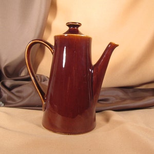 May include: A brown ceramic coffee pot with a lid and a handle. The pot is sitting on a brown surface.