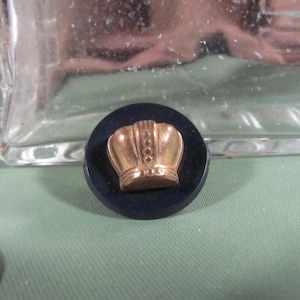 May include: A gold crown button with a blue background. The crown has a detailed design with a raised center and a textured surface.
