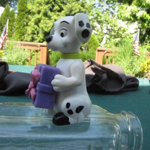 Może przedstawiać: Figurkę białego i czarnego dalmatyńczyka trzymającego fioletowe pudełko prezentowe z różową kokardką. Figurka stoi na przezroczystej szklanej powierzchni.