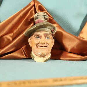 May include: A ceramic wall plaque of a man's head wearing a green hat with a red and white flower. The man has a white beard and mustache and is smiling. The plaque is decorated with a small, white column on top.