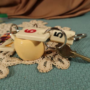 May include: A set of keys with a cream-colored ball charm and two white plastic tags with the numbers "6" and "5" on them. The keys are attached to a silver key ring.