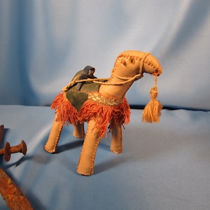 May include: A tan camel figurine with a red and green saddle and a small blue rider. The camel has a long neck and a tassel on its head.