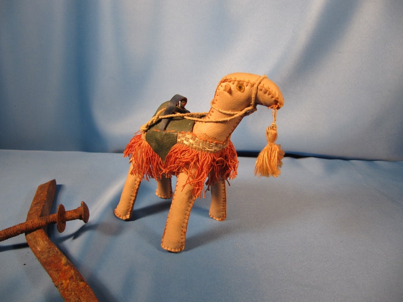 May include: A tan camel figurine with a red and green saddle and a small blue rider. The camel has a long neck and a tassel on its head.