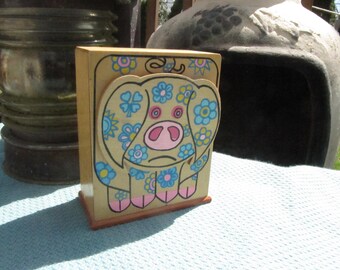 May include: A wooden piggy bank with a whimsical design. The pig is painted with a light brown base and decorated with blue and pink flowers. The piggy bank has a slot on the top for coins.