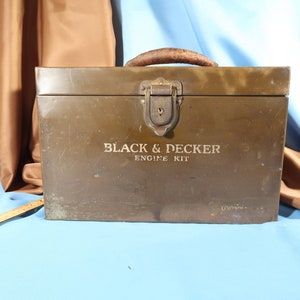 Op de afbeelding: Een vintage groene metalen gereedschapskist met een leren handvat en een messing sluiting. Op de voorkant van de gereedschapskist staat "BLACK & DECKER ENGINE KIT" gedrukt.