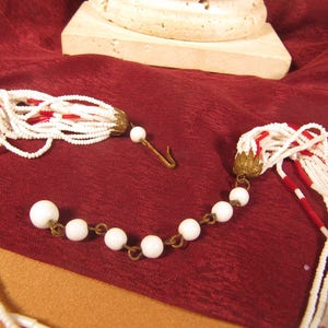 May include: A white and red beaded necklace with a tassel on each end. The necklace is made of small, round beads and has a gold-toned clasp.