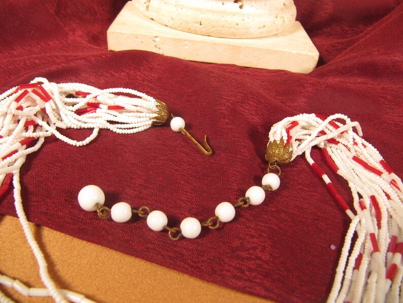 May include: A white and red beaded necklace with a tassel on each end. The necklace is made of small, round beads and has a gold-toned clasp.