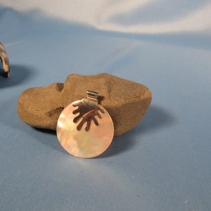 May include: A round, iridescent, mother-of-pearl pendant with a silver-toned, hand-shaped cutout design. The pendant is hanging from a silver-toned bail.
