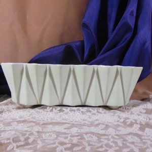 May include: A white ceramic planter with a geometric design. The planter has a rectangular shape with a series of triangular indentations along the sides. The planter is sitting on a white lace tablecloth.