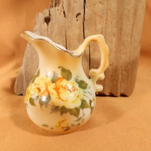 May include: A small, cream-coloured ceramic jug with a floral design. The jug has a scalloped rim and a handle with a floral design. The flowers are yellow and white with green leaves.