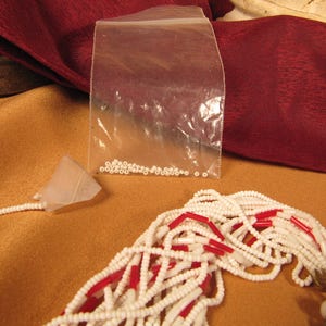 May include: A white and red beaded necklace with a gold clasp. The beads are small and round, and the necklace is very long. There is a small bag of white beads in the background.