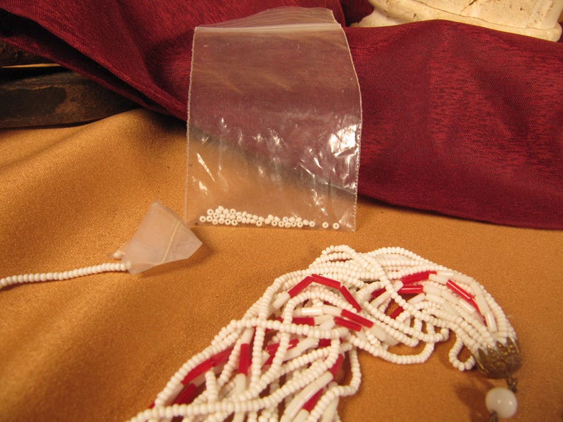 May include: A white and red beaded necklace with a gold clasp. The beads are small and round, and the necklace is very long. There is a small bag of white beads in the background.