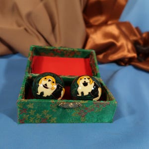 May include: A pair of decorative dog-themed spheres, each featuring a smiling dog design in yellow, black, and white, are displayed inside an open, green velvet box with a red interior. The box has a decorative clasp and is set against a blue backdrop.