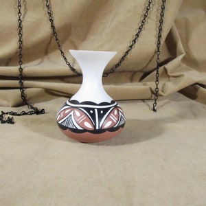 May include: A white ceramic vase with a brown, black, and white geometric design. The vase is sitting on a brown fabric surface.