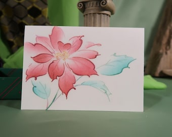 May include: A watercolor painting of a poinsettia flower with red petals and yellow center. The flower has green leaves and is on a white card. The card is on a green surface.