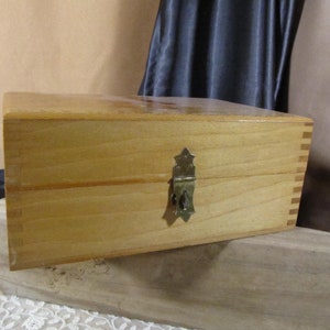 Puede incluir: Una caja de madera con un cierre de latón. La caja está hecha de madera clara y tiene un diseño sencillo.
