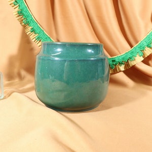 May include: A green ceramic vase with a rounded shape and a smooth, glossy finish. The vase is sitting on a light brown fabric surface.