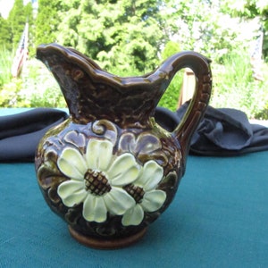 May include: A brown ceramic jug with a raised floral design. The jug has a handle and a wide mouth. The flowers are white with yellow centres.