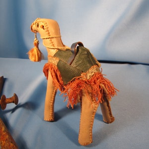 May include: A small, tan leather camel figurine with a green leather saddle and orange fringe. The camel has a long neck and a small head with a tassel.
