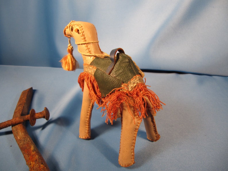 May include: A small, tan leather camel figurine with a green leather saddle and orange fringe. The camel has a long neck and a small head with a tassel.