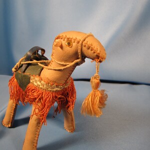 May include: A tan leather camel figurine with a blue saddle, orange fringe, and a tan tassel. The camel has glass eyes.