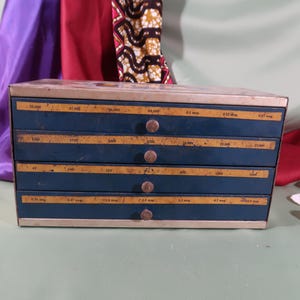 May include: A rectangular metal storage cabinet with four horizontal drawers. Each drawer front is dark blue with a horizontal tan label strip featuring printed numerical data and text. Each drawer has a small, centered, circular metal knob.