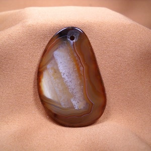 May include: A brown and white agate pendant with a hole for a cord or chain. The pendant is oval shaped and has a natural, polished finish.