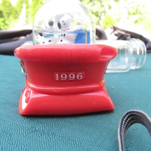 Może przedstawiać: Czerwono-biała kula śnieżna z dwoma szczeniakami dalmatyńczyka w środku. Kula śnieżna stoi na czerwonej podstawie z nadrukowanym rokiem 1996.