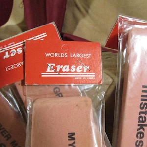 May include: Three pink erasers packaged in plastic. The erasers are labeled "Worlds Largest Eraser" and "My Grandpa Never Makes Mistakes".